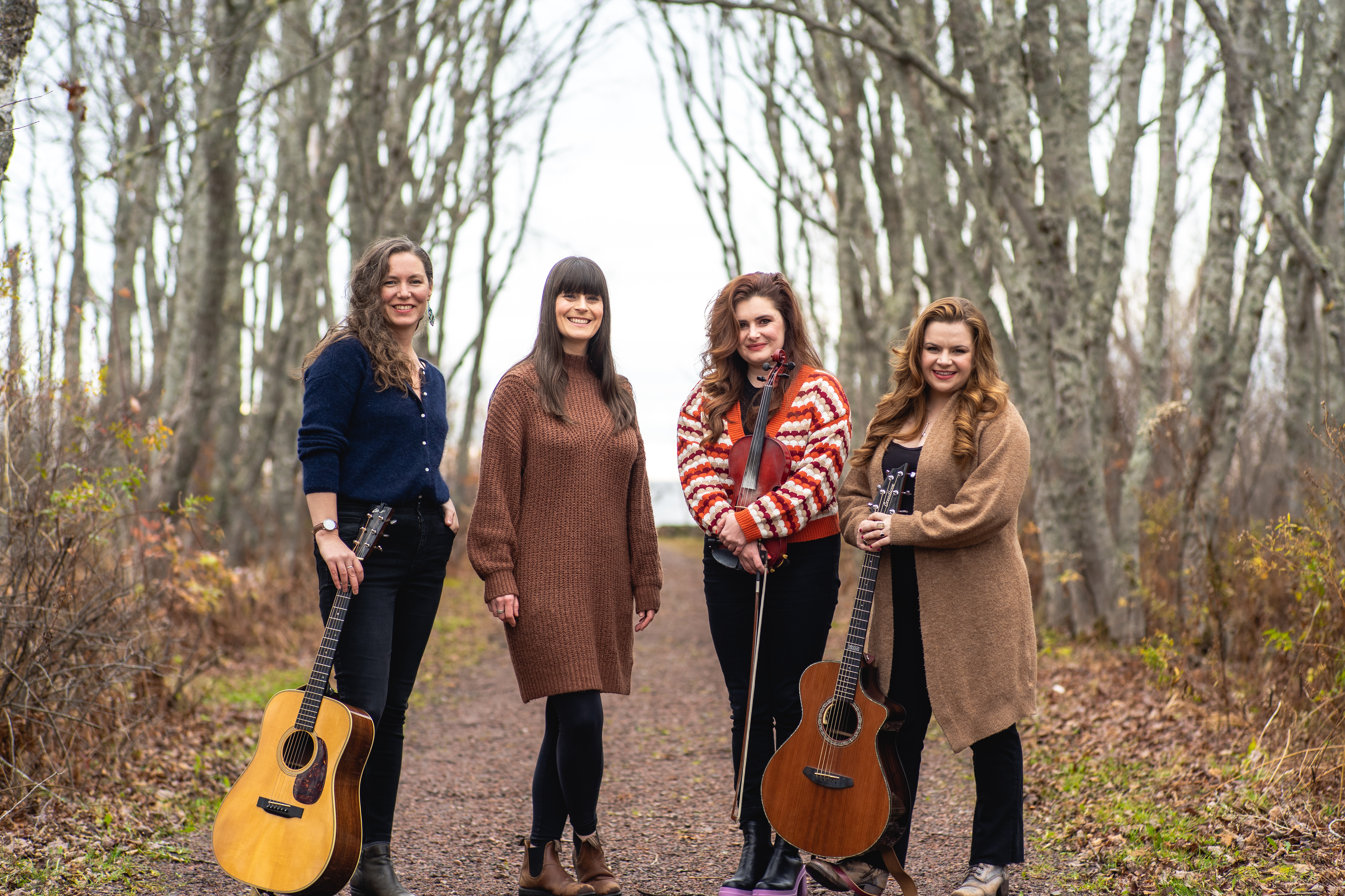 4 women standing on a path outside, two hold guitars, one is holding a fiddle