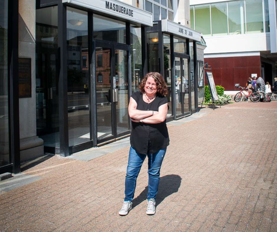 Melissa Cameron, curly brown hair, stands out front of the entrance to the Playhouse Theatre on a sunny day. She smiles with her arms crossed, wearing a black t-shirt and jeans. 