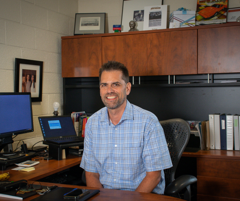 Tim Yerxa, short black hair and a short sleeve blue dress shirt, sits behind his office desk smiling. 