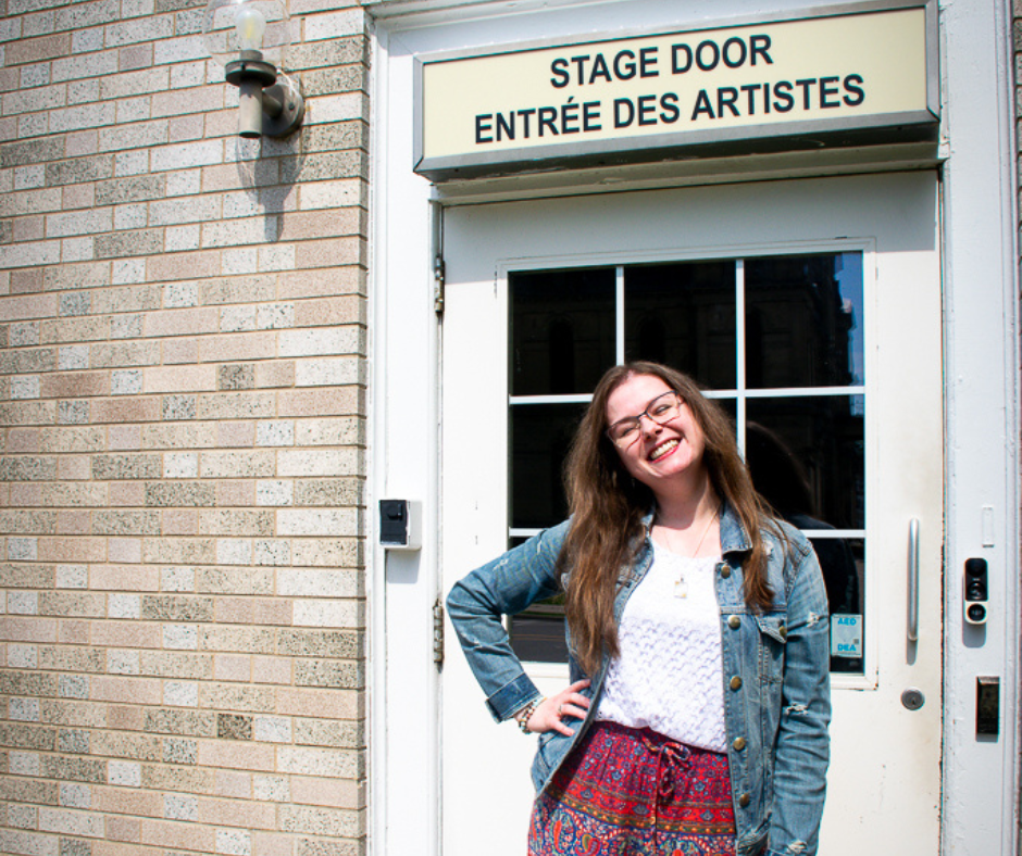 Katelyn Goodwin stands in front of the stage door entrance of the Fredericton Playhouse. Her long brown hair is being hit by the sun, she stands smiling while wearing a jean jacket and colourful red pants. 