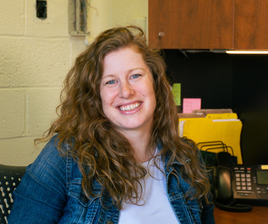 Meghan Callaghan, curly red hair, sits at her desk smiling. She is wearing a denim jacket and a white t-shirt. 