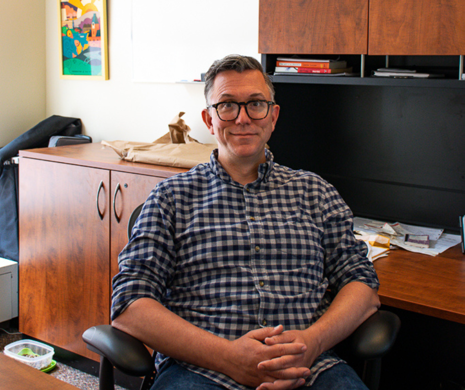 Jeff Richardson, dark hair and glasses, smiles in his office wearing a blue checkered dress shirt and jeans.