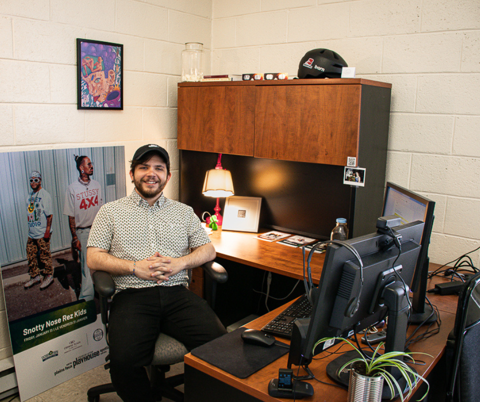 Jason McIntyre sits behind his office desk with a black ball cap on and a white and green button up shirt. He has his fingers interlocked and is smiling at the camera. 