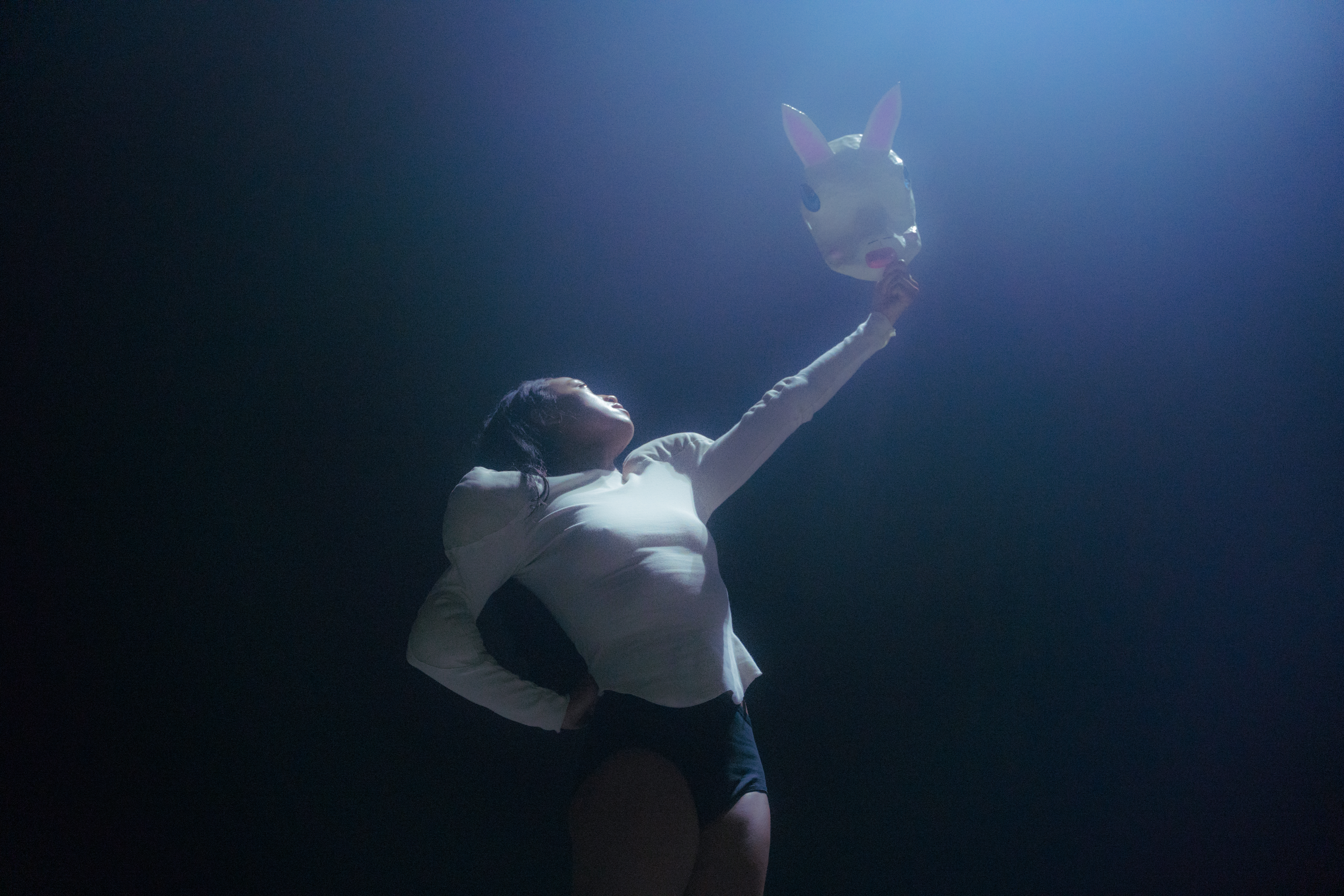 A dancer stands under a spotlight holding a white rabbit mask up in the air.