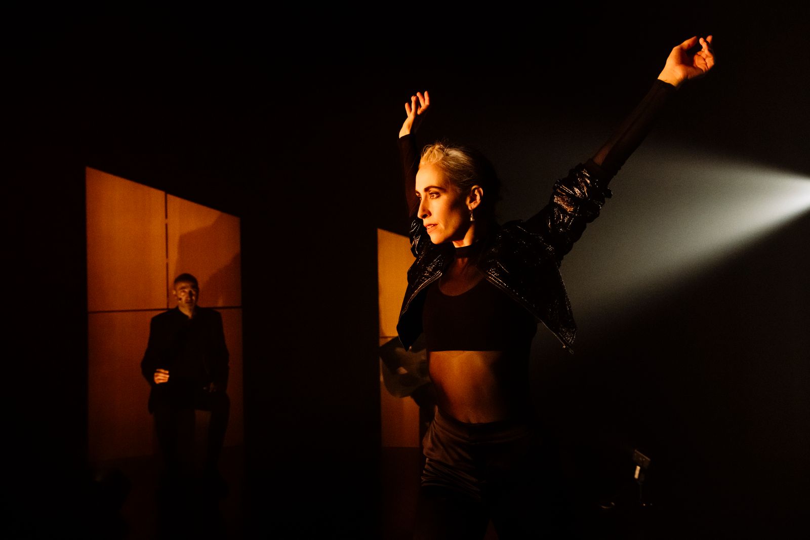 A dancer strikes a bold pose with her arms spread wide above her, dressed in fashionable black dance clothing in a smoky looking, orange-lit room.