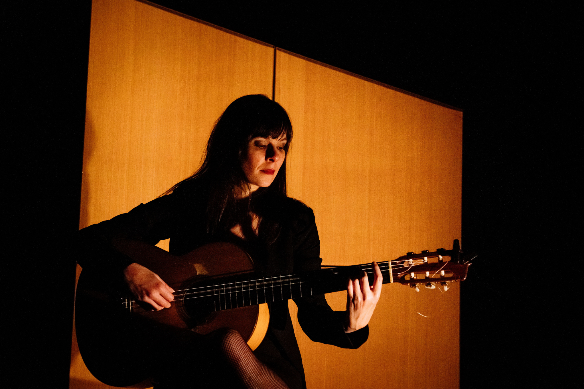 A black haired woman sits in front of a moody looking orange background playing a classical guitar with a focussed look on her face.