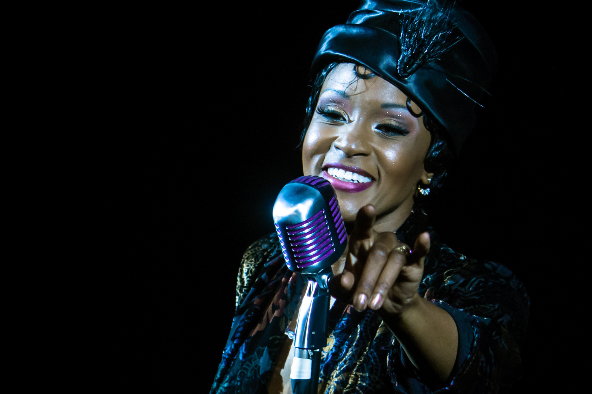 An actress stands behind a microphone smiling, dressed as Josephine Baker (a stylish silk robe and matching hat that showcase the style of early cabaret performances).