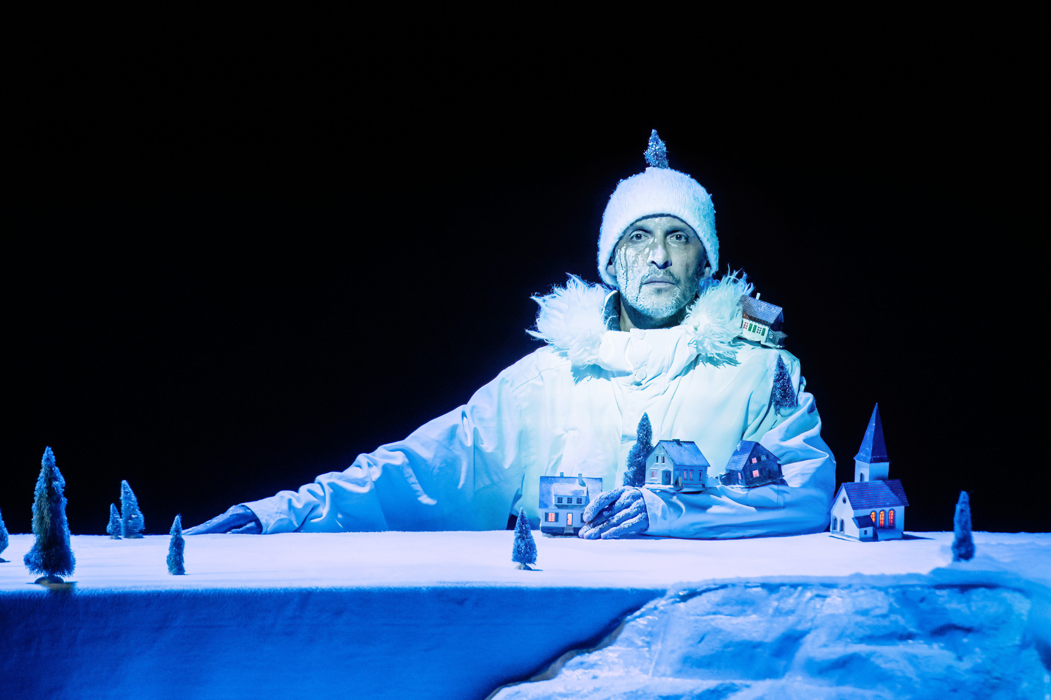 A man in a white winter jacket stands behind a miniature scene of a snowy wooden forest. He is wearing all white, giving the illusion that he himself is a snowy mountain within this landscape.