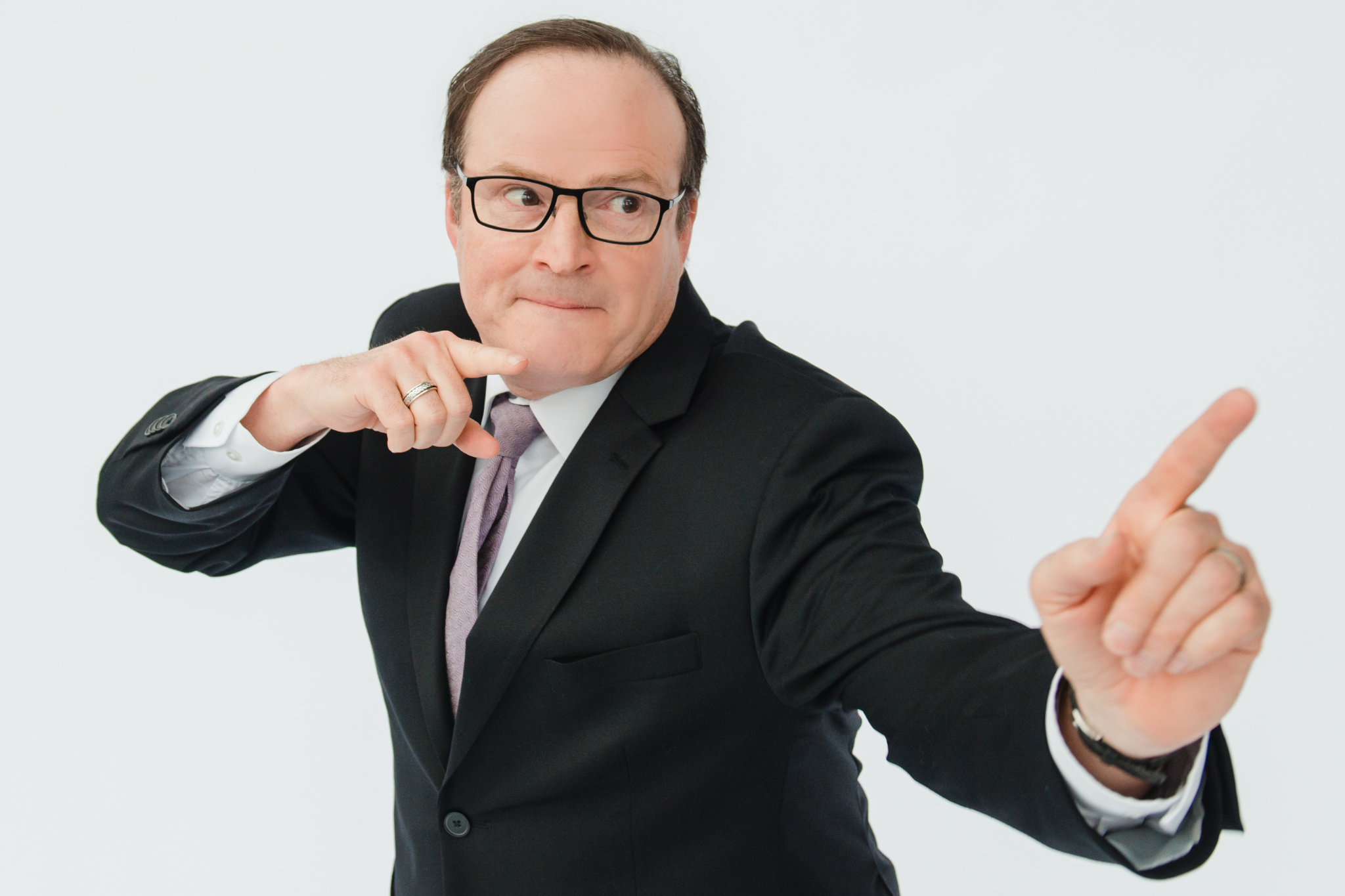 Steve Patterson stands with his fingers pointed in front of him, pulling a funny face while wearing a black suit. 