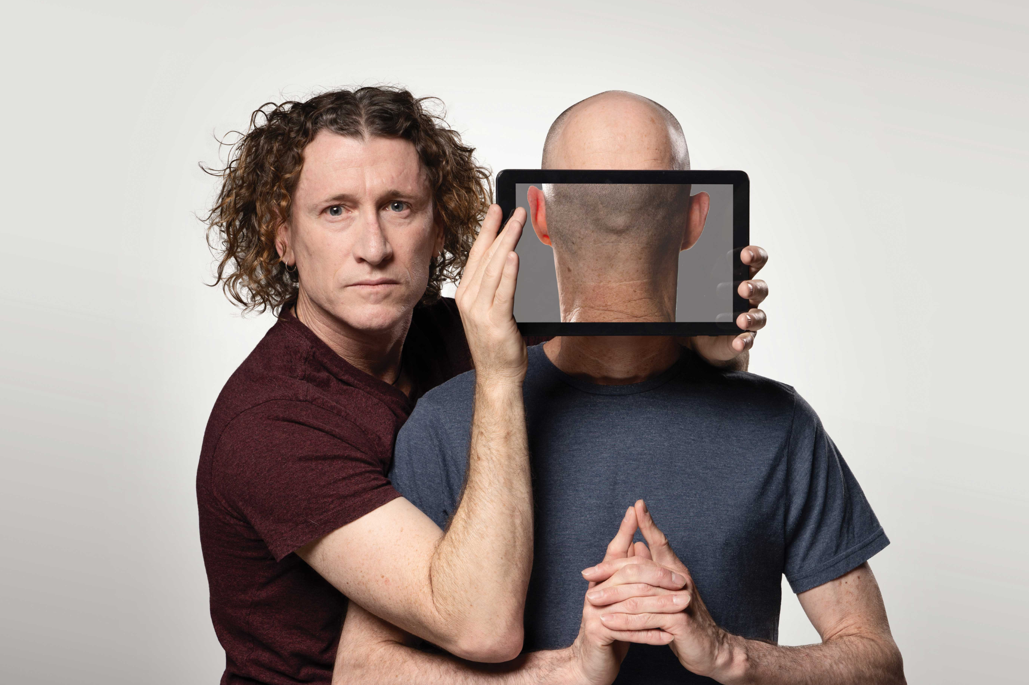 The two brothers stand facing the camera, while one brother holds an iPad in front of the others face. On the iPad is an image of the back of his brother's head, giving the image a strange and humorous quality. 