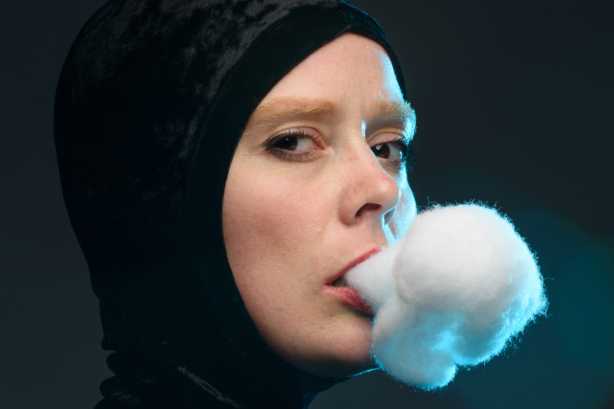 A woman wearing a tight fitting black hood stands in front of a smoky blue background, while a large cloud shaped cotton ball hangs from her mouth giving the illusion that she is smoking. 