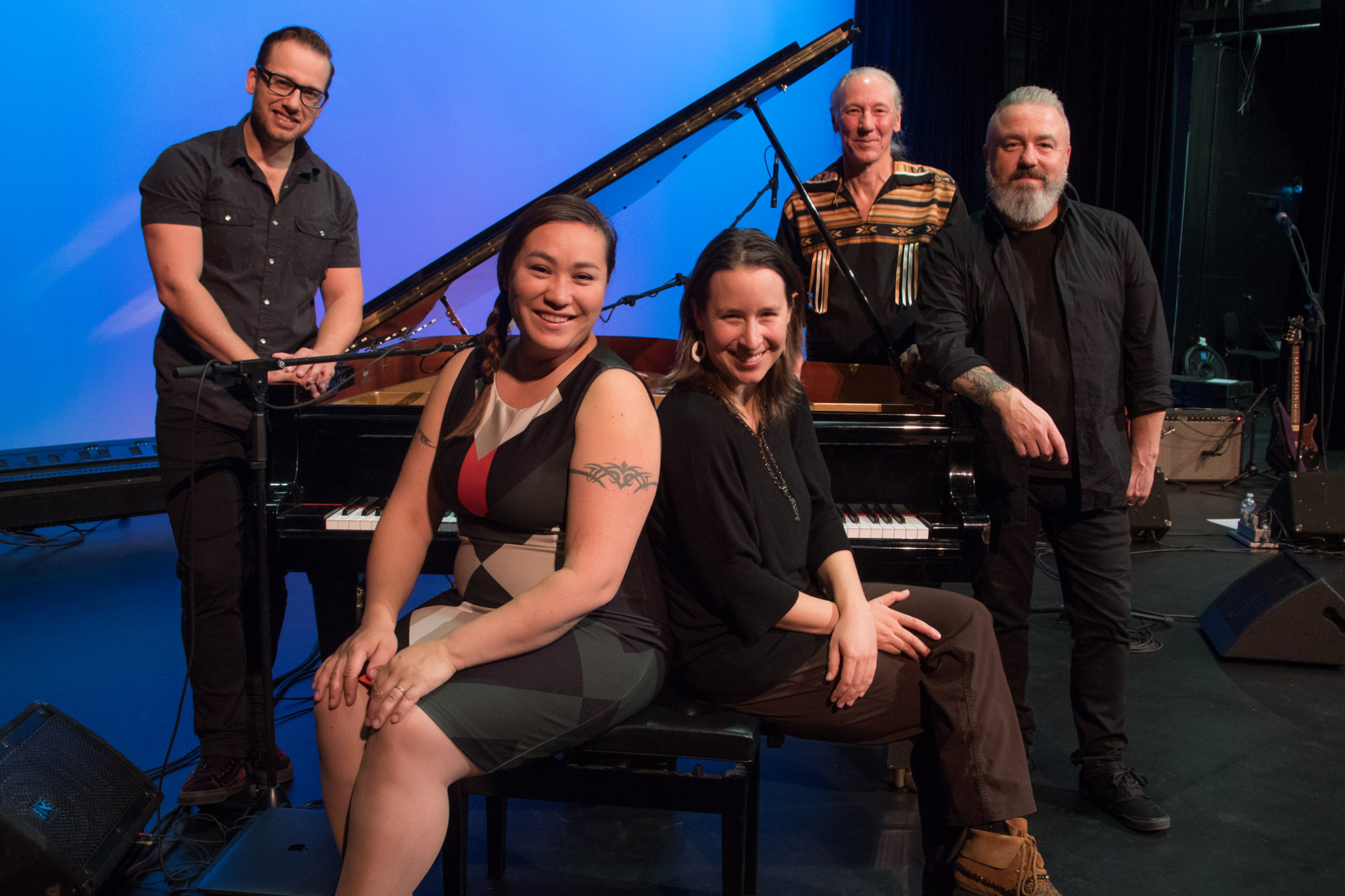 A group of five musicians stand around a piano and smile at the camea. Several are wearing First Nations inspired clothing.