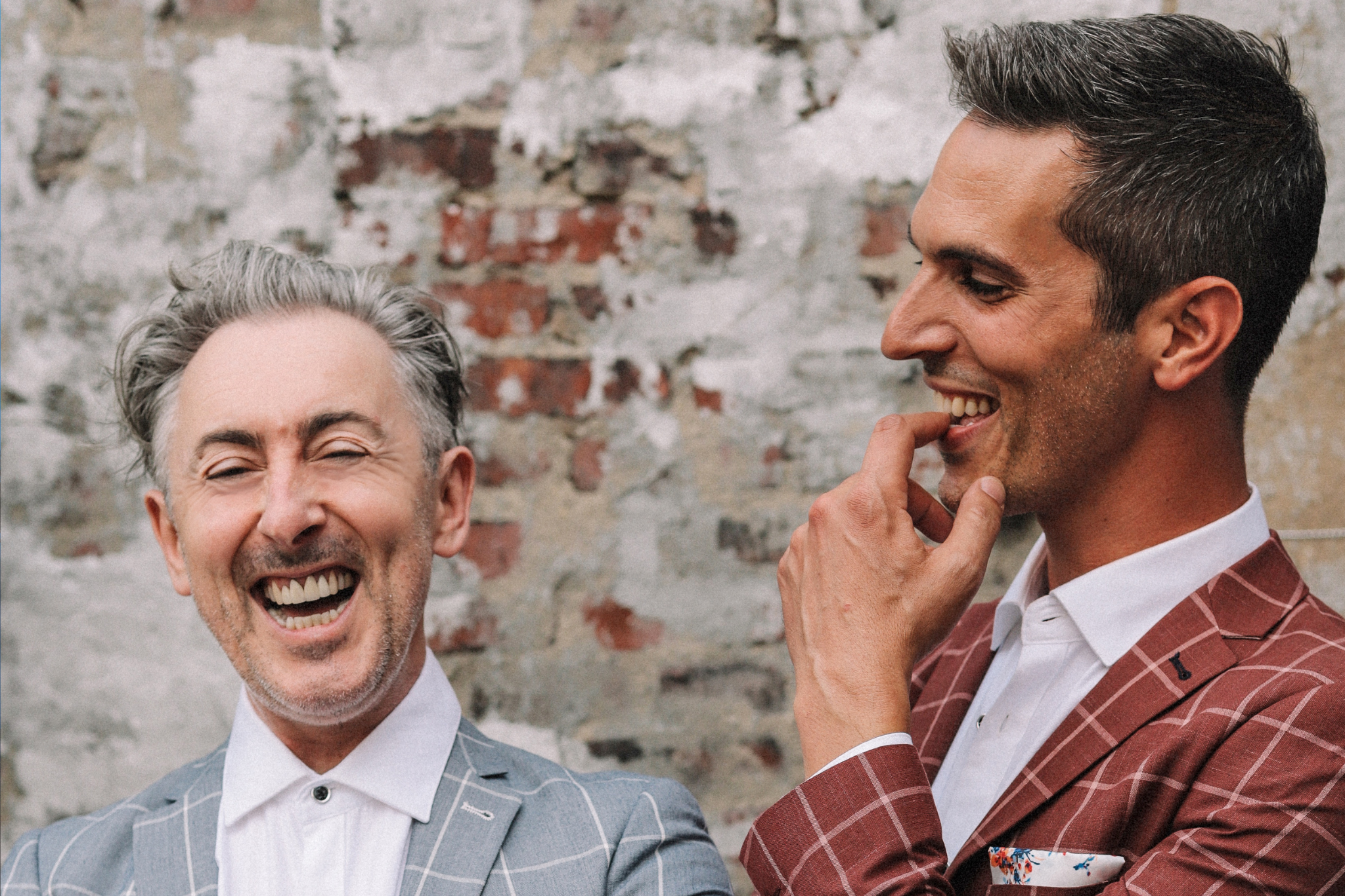 Alan Cumming and Ari Shapiro stand in front of a brick wall, each wearing sharp looking stripped suits. Alan has his eyes closed in laughter as Ari looks on at him admiringly. 