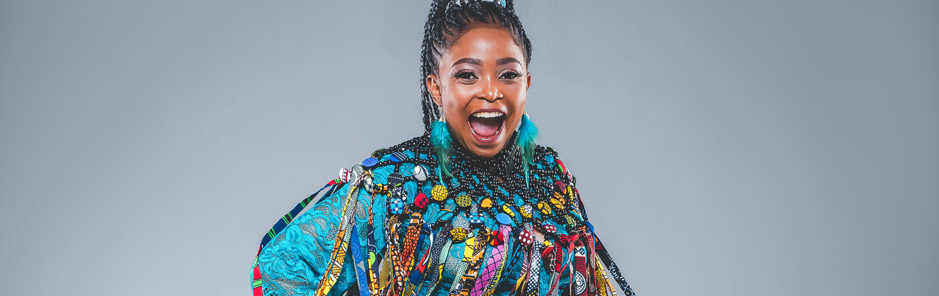 A close up of Nomfusi in a light blue tassled outfit, pictured from the chest up. She is looking right at the camera with her mouth open in a big, energetic smile