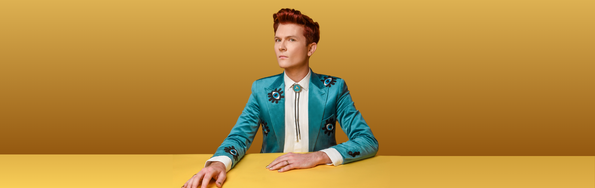 Rhys Nicholson sitting at a yellow table surface against a darker yellow background. They are wearing a light teal blue jacket and a white shirt, looking at the camera with their head turned slightly sideways