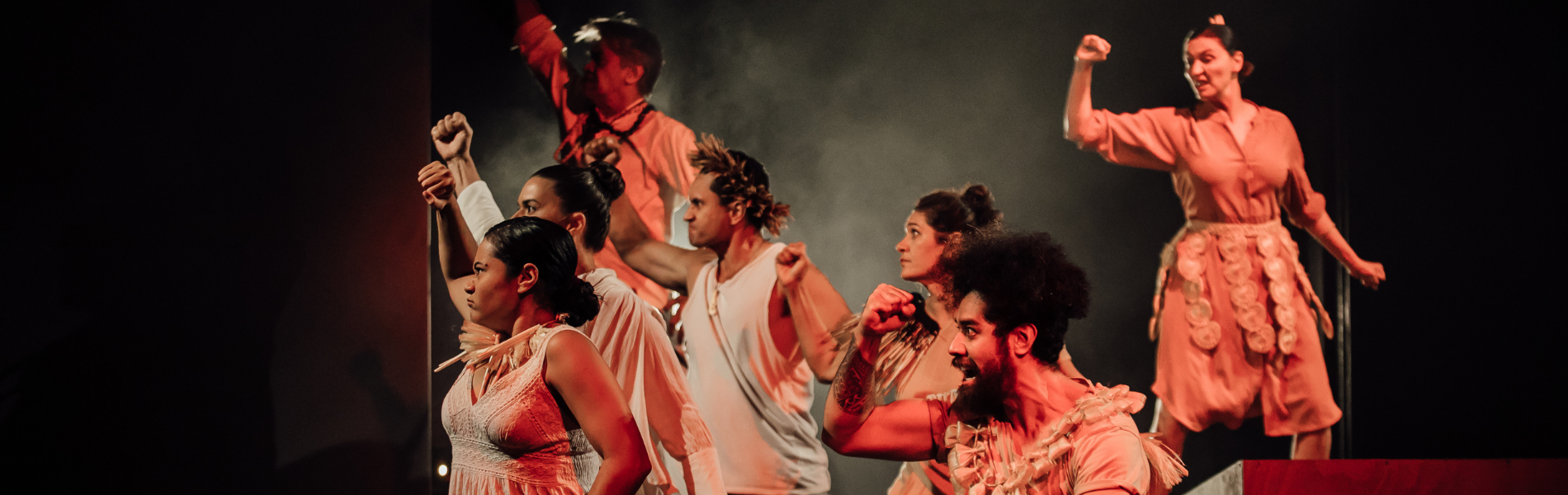 A group of actors from the UPU Collective standing against a dark background, all looking to the left of the camera, arms raised protectively