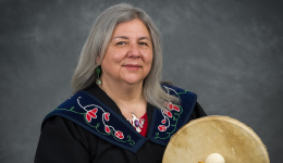 A photo of Lisa Perley Dutcher in a black shirt, holding a drum, against a grey backdrop