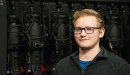 Finn Boehm in a black collared shirt standing in front of a background of stage lights hanging on the wall