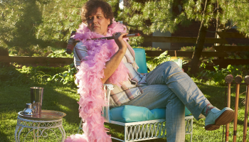 Ron Sexsmith with a pink feather boa sitting on a chair among trees