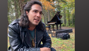Jeremy Dutcher outside in nature with a piano on a stage in the background.
