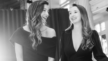 Two women smiling at each other, standing beside a piano.