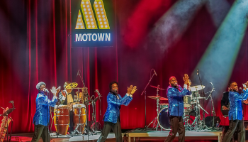 5 men in blue suit coats dancing in sync with musicians behind them and a sign that says Motown.