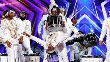 A group of drummers and dancers dressed in all white against a purple background
