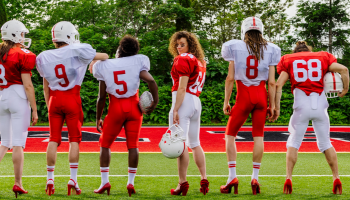 A row of people facing away wearing red and white football uniforms and red high heels, the person in the centre is looking back at the camera