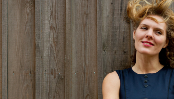 A cropped picture of a woman in a blue sleeveless top standing against a fence, her hair blowing in the wind
