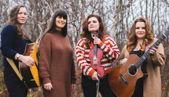 Four women holding instruments, standing together in front of bare trees