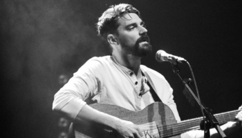 A black and white image of Tim Baker in the spotlight, singing and playing guitar.