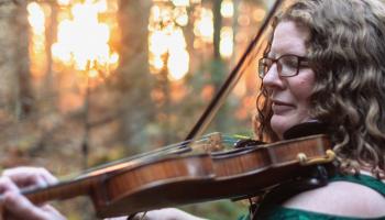 Katherine Moller playing the fiddle outside with trees and a sunset in the background.