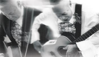 A blurred black and white image of Tommy Emmanuel, CGP playing guitar.
