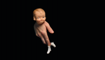 A doll head at the top of a person's hand, shaped to look like the body and legs of the doll with tiny skates on the end of the fingers, against a black backdrop