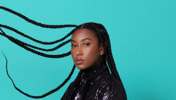 Dominique Fils-Aimé in front of a teal blue background with her hair in braids thrown out to the side of the photo