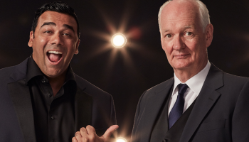 Asad Mecci and Colin Mochrie in black suit jackets standing against a black background with stage spotlights