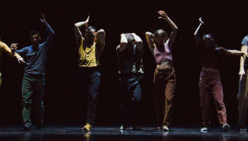 7 contemporary dancers in a line, darkly lit against a black background