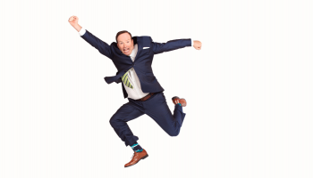 Steve Patterson jumping in the air, arms outstretched at different angles, and legs bent. He's wearing a full suit and is in front of a flat white background