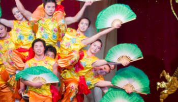 The Peking Acrobats holding large green fans out to the side as all 10 balance on a single bike