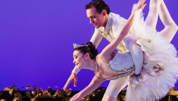 Two ballet dancers in front of a purple background, one in a pink tutu, being held forward by the other one in a suit.