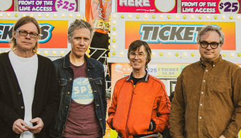 The 4 members of Trans-Canada Highwaymen looking at the camera, standing in front of a colourful carnival ticket booth