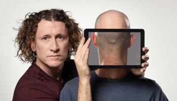 One man standing next to another holding a screen in front of the other's face. On the screen the image looks like the back of the man's head.