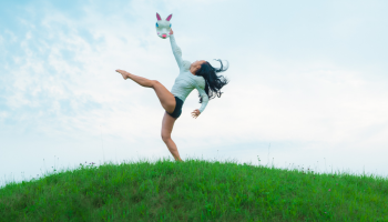 A dancer striking a post with limbs extended in front of a cloudy blue sky, on top of a bright green hill