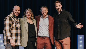 Four comedians posing on a stage in front of a lit sign.