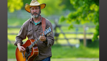 An image of Tom Green playing a guitar in a field of green.