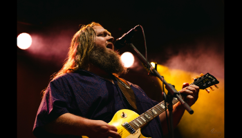 An image of Matt Andersen singing into a mic while playing guitar with pink and yellow lighting behind. 