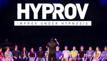 A group of people in chairs on the stage with the words, 'HYPROV Improv under hypnosis' in white lettering above them.