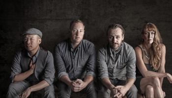 The four band members sit against a wooden wall.