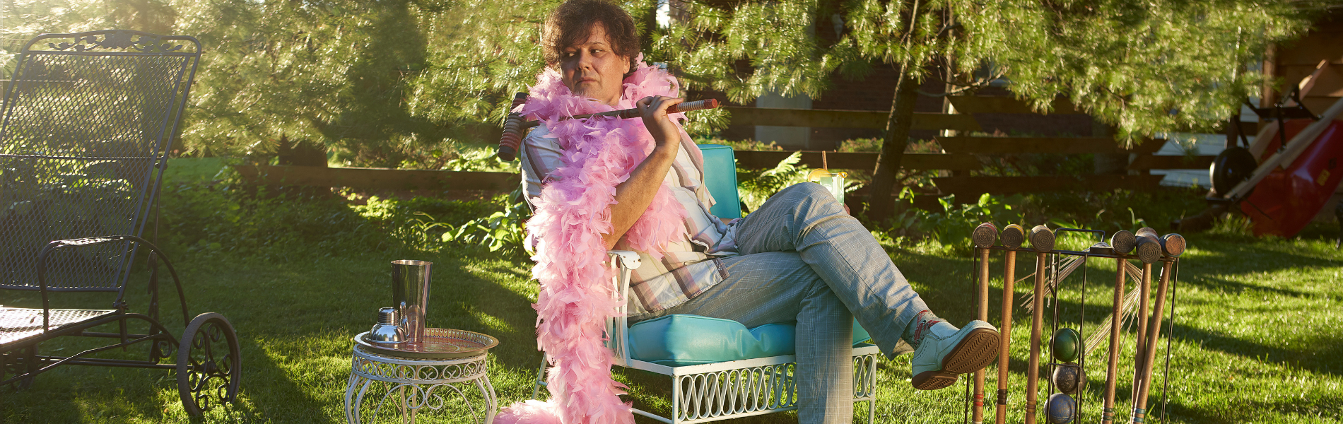 Ron Sexsmith with a pink feather boa sitting on a chair among trees