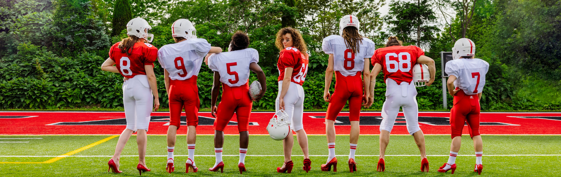 A row of people facing away wearing red and white football uniforms and red high heels, the person in the centre is looking back at the camera